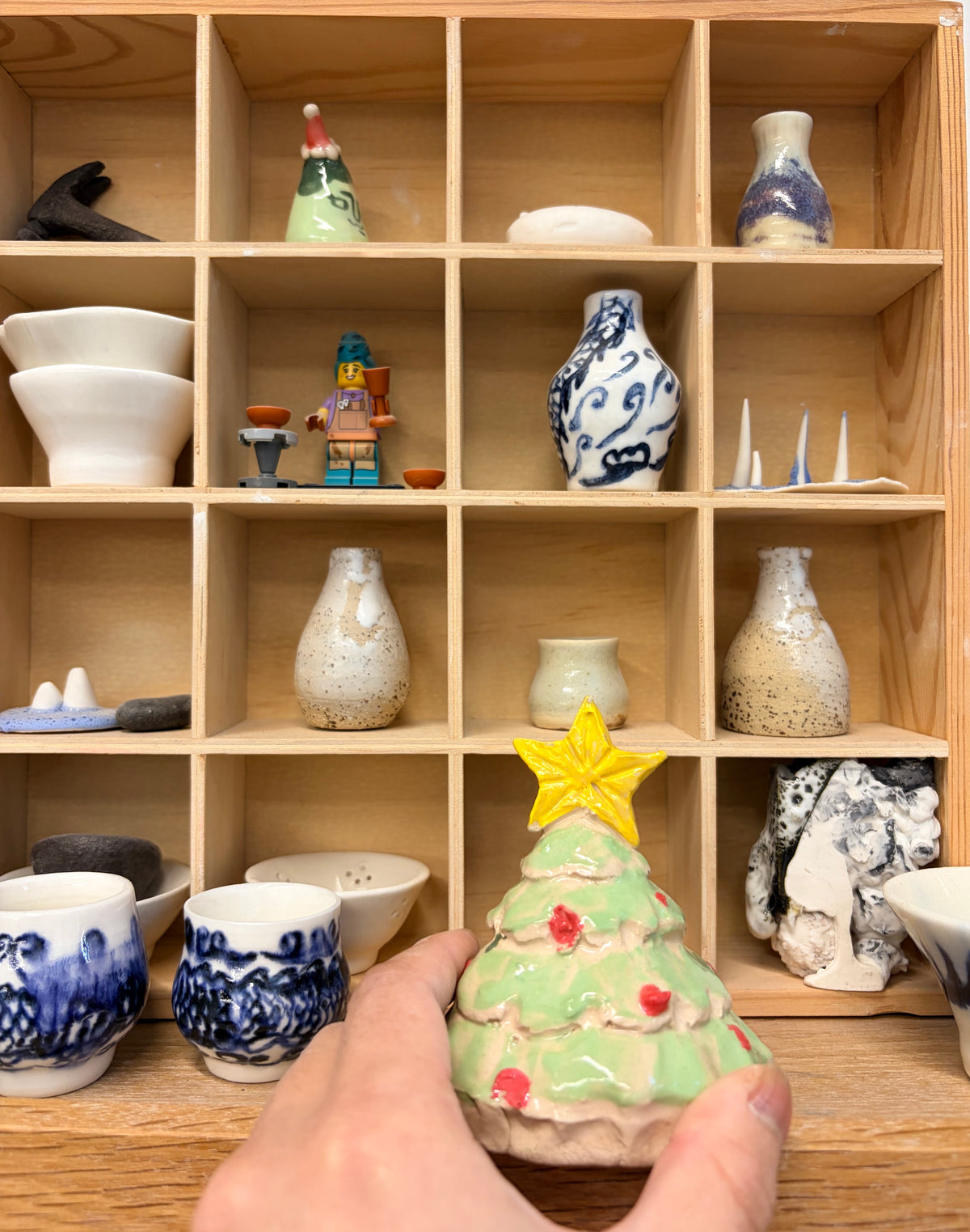 A hand holds a small ceramic Christmas tree ornament with a yellow star topper, in front of a wooden display shelf filled with other handmade ceramic items.