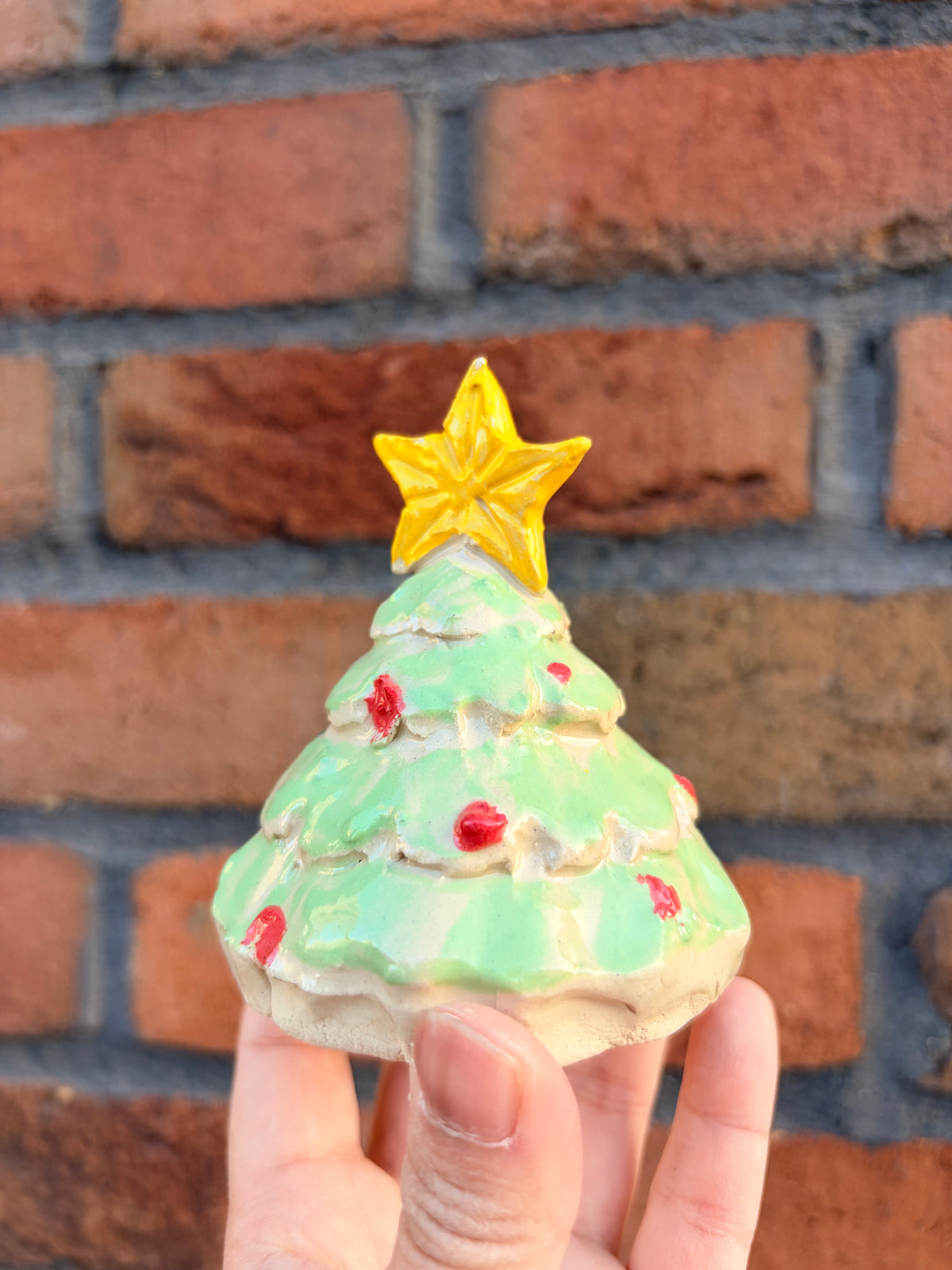 A hand holding a small ceramic Christmas tree ornament with a light green body, red ornaments, and a yellow star topper, set against a blurred red brick wall background.