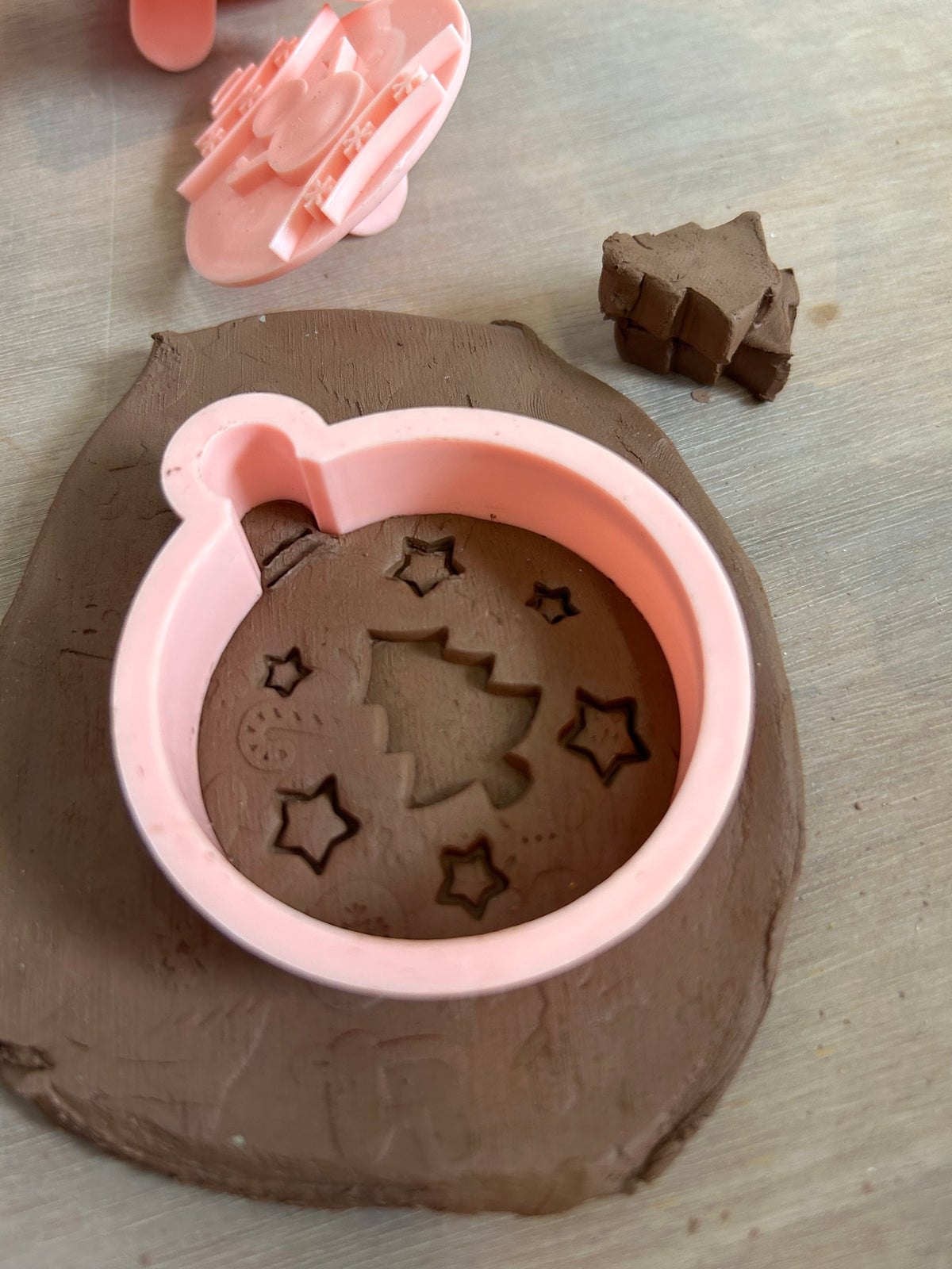 A pink plastic clay cutter with a Christmas tree and star design, shown pressed into brown modeling clay on a light wood surface, with the matching pink cutter and a small cut piece of clay in the background.